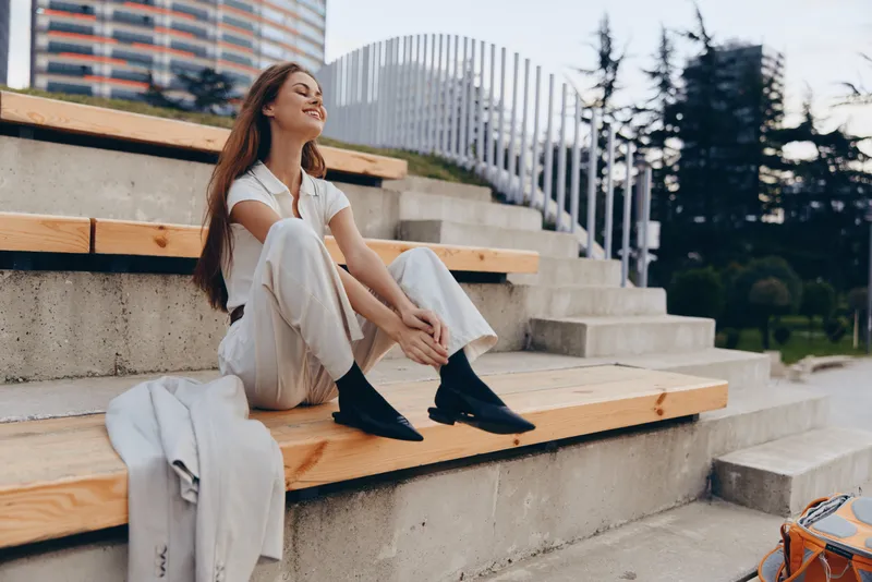 Young woman in casual white outfit sitting relaxed on wooden steps outdoors with serene expression, urban background and soft natural light, fashion lifestyle concept