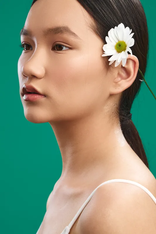 asian woman with a delicate flower tucked behind the ear gazes softly forward, a minimalist portrait against a vivid green backdrop, with natural light enhancing smooth skin and a calm, graceful aura.