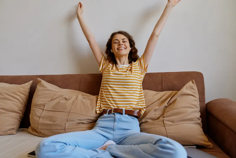 Young woman with striped shirt raises arms in celebration on a cozy living room couch as she sits cross legged on cushions, enjoying a relaxed happy moment at home.