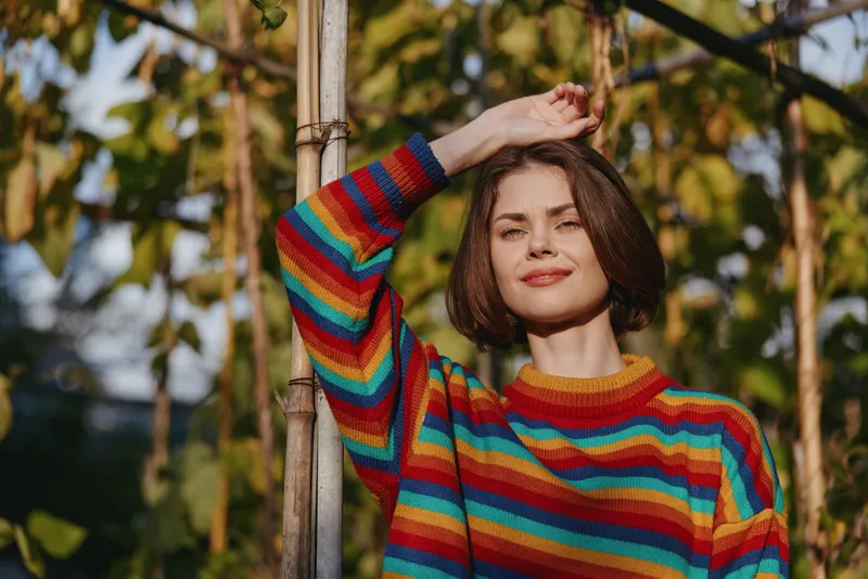 Young woman in her 20s wearing a colorful striped sweater, outdoor portrait with short brown hair, smiling and leaning on a bamboo pole in warm sunlight, casual lifestyle.