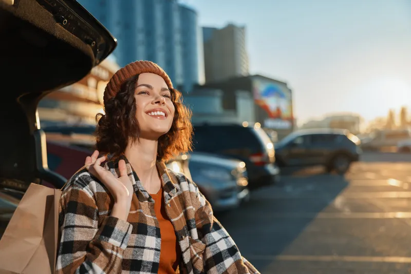 Woman smile by car trunk in parking lot at sunset city golden hour, beanie and plaid jacket, candid lifestyle portrait with authenticity, mindful living and emotional storytelling vibe.