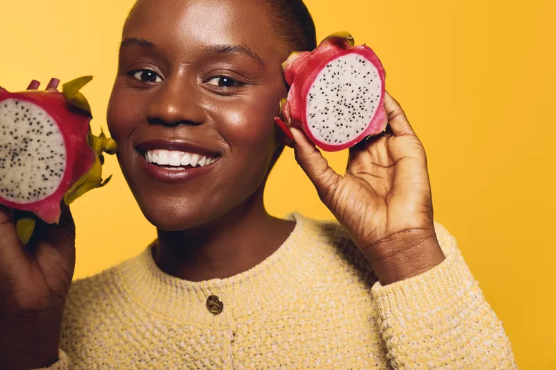 Smiling dark skinned African woman holds two halves of dragon fruit close to her face on yellow background. Concept of healthy lifestyle, exotic fruit, wellness, and natural beauty.