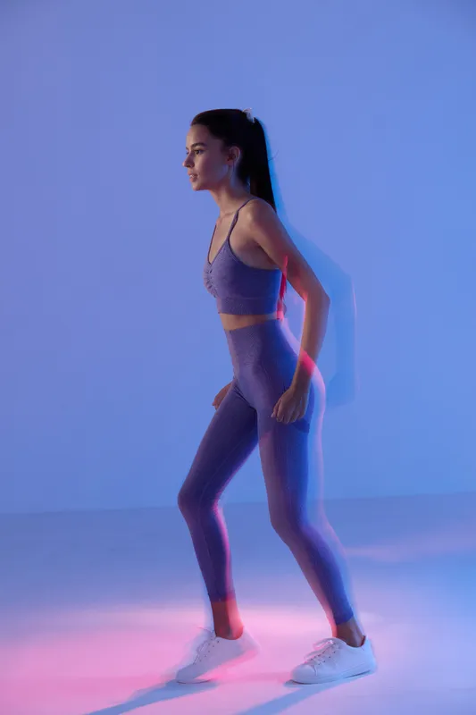 Woman sport fitness wearing purple activewear, poised in a dynamic pose with a focused expression, illuminated by soft purple and pink neon lighting in studio.