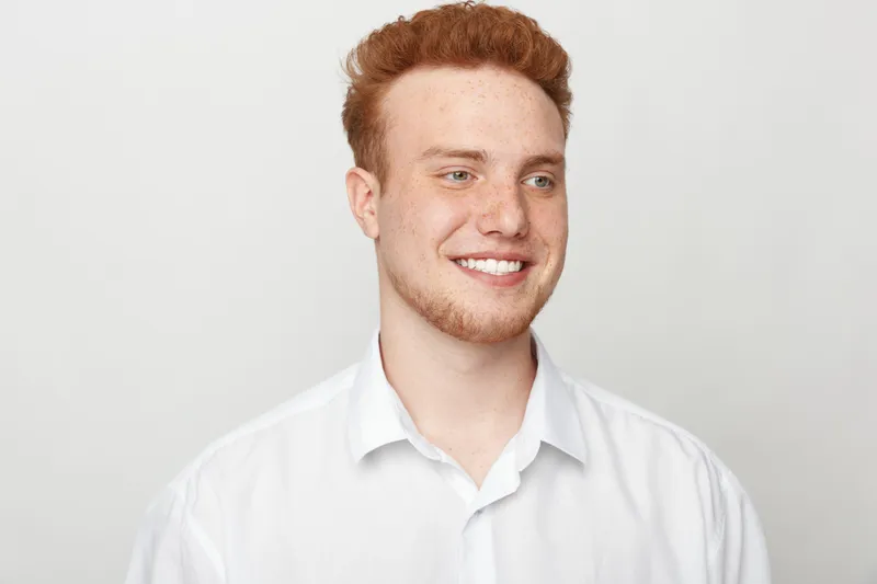 Smiling young man with red hair and beard wearing white shirt looks away on light background. Concept of happiness, casual style, youth and positive emotions in studio portrait.