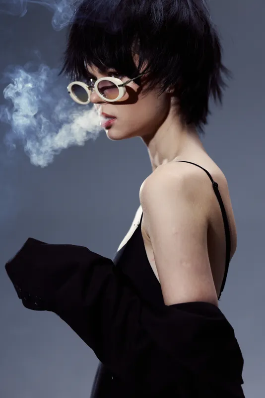 Stylish woman with short hair wearing sunglasses and smoking, dressed in black with moody lighting and smoke detail