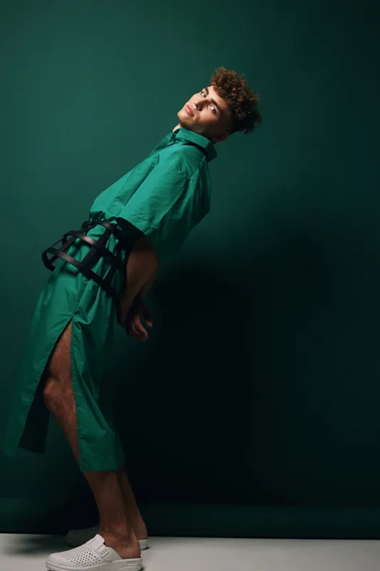 Young man in green outfit and white shoes posing with hands behind back against dark green background in studio for fashion and style concept.