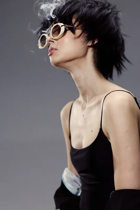 Stylish young woman wearing round sunglasses with short black hair and smoke against gray background, expressing cool attitude.