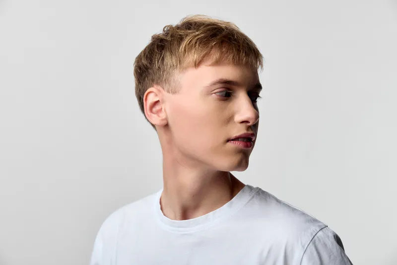 Young man, profile view, calm expression and clean background, white shirt, studio lighting highlights facial features, minimal setting, modern lifestyle photo suitable for fashion, editorial, or