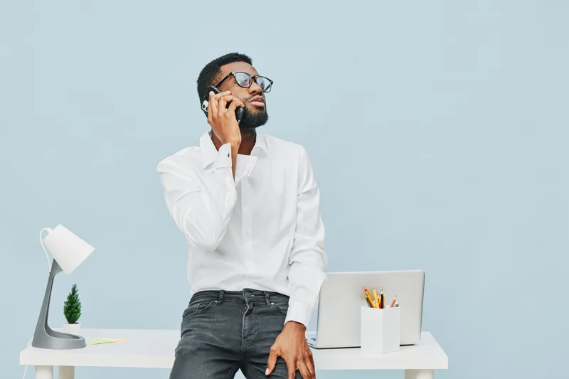 Modern busy man speaking on cellphone while standing in office, wearing white shirt and black jeans, with laptop and stationery on desk, minimalistic workspace background. People lifestyle concept, co