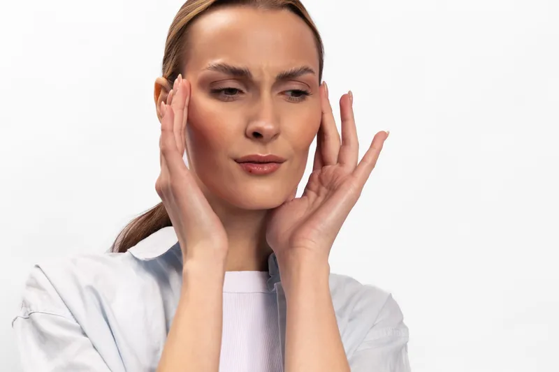 Young woman with fair skin and light hair touches her temples with both hands, showing a tense expression. Concept for headache, stress, pain, and discomfort in health and wellness.