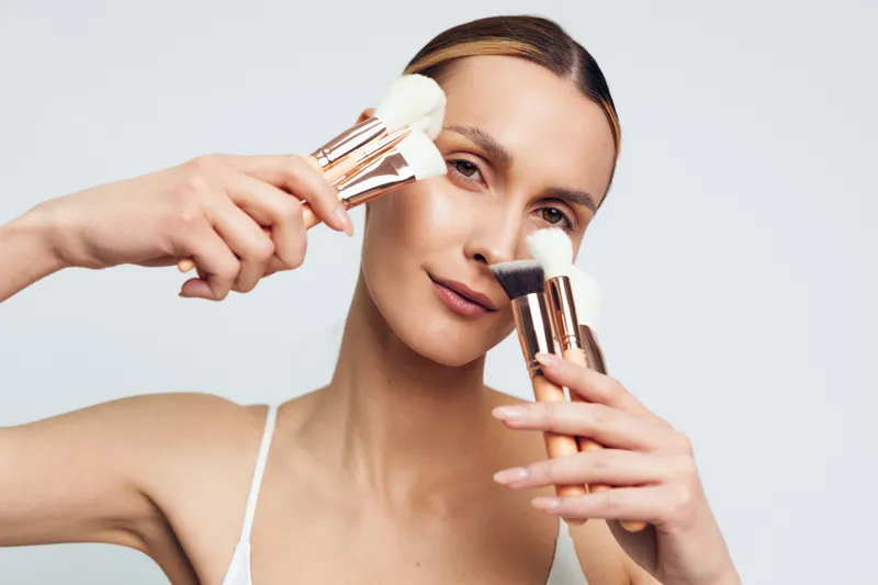 Youthful woman with dark hair uses multiple makeup brushes to blend powder on her face in a bright studio, presenting a calm focused mood and professional beauty routine