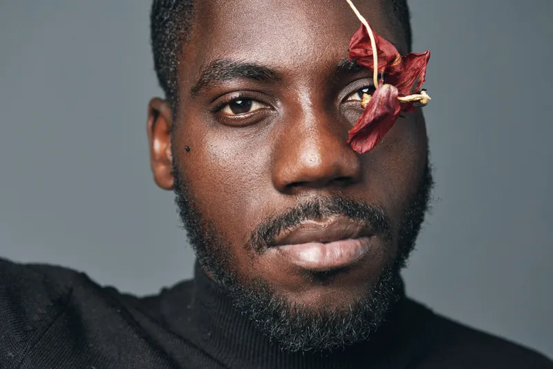 African man portrait with dried flower on face in creative conceptual visual style. Closeup of black male model with beard and serious expression on gray background for artistic photography.