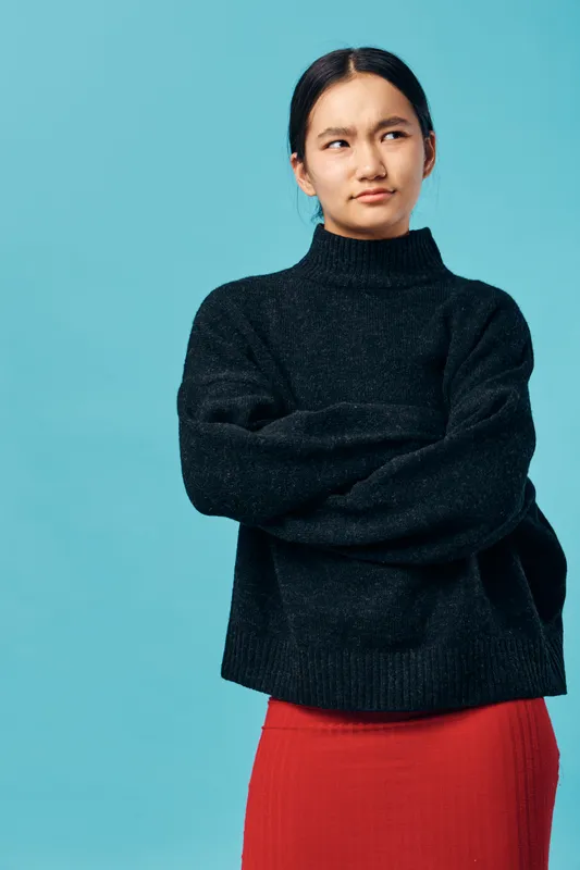 Woman with crossed arms wearing black sweater and red skirt stands against blue background with skeptical expression looking sideways in casual style portrait for fashion and attitude.