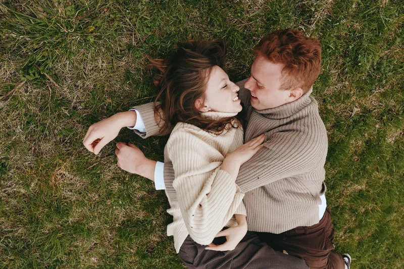 A couple lies on grass embracing softly, smiling and connected, wearing cozy sweaters. Natural light and outdoor setting convey warmth, affection, and relaxed lifestyle moment.
