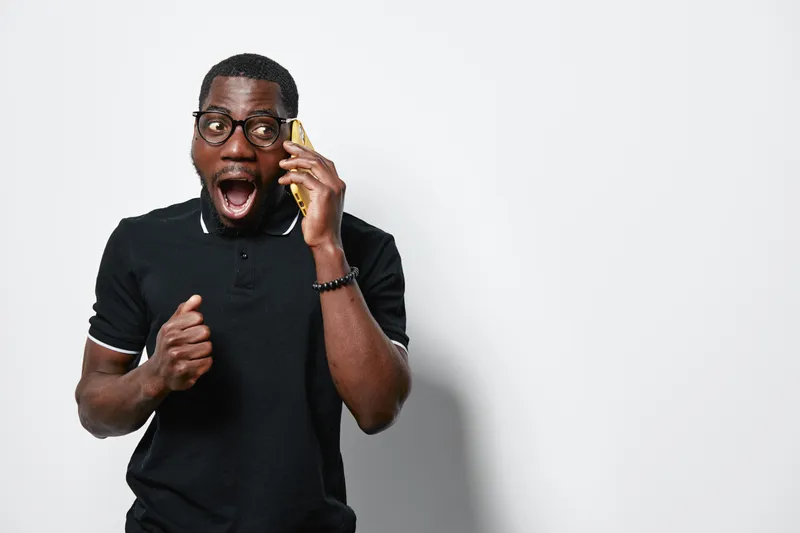 African American man with dark skin and glasses wearing black polo shirt talking on smartphone with surprised expression on white background. Communication, technology and emotion.