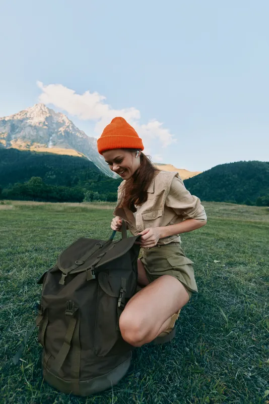 Young woman outdoors kneels beside a large backpack in a green meadow with distant mountains, smiling softly as she begins unpacking gear for a day in nature.