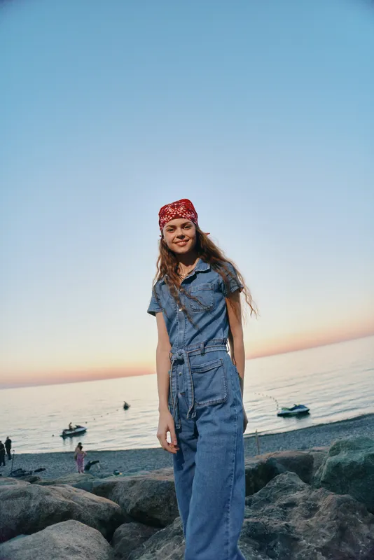 lifestyle woman in modern boho-western denim street style enjoys seaside sunset with relaxed smile, casual outfit, and film color tones enhancing warm mood