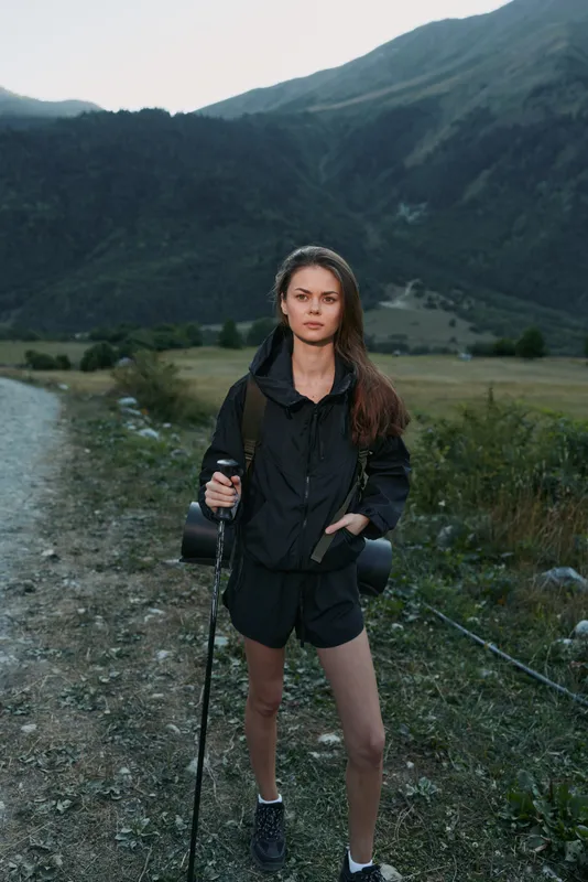 A lone hiker stands on a gravel trail in a mountainous landscape, wearing a dark rain jacket and shorts, with a backpack and trekking pole, ready for outdoor adventure.