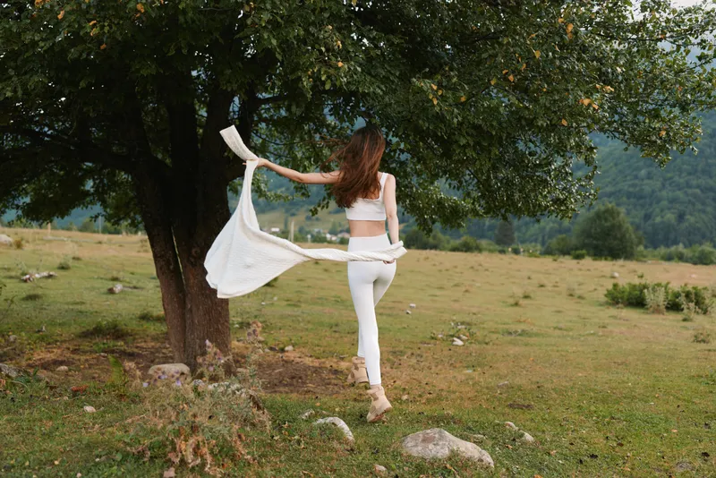 Young woman in white stands in a grassy field, scarf fluttering in the breeze, enjoying outdoor moments. Natural light creates a carefree mood of nature and wellness.