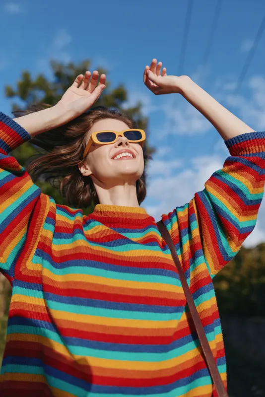 Woman wearing rainbow sweater and yellow sunglasses smiling outdoors under a sunny sky, joyful young adult in casual outfit raising arms, lifestyle portrait for travel and fashion.