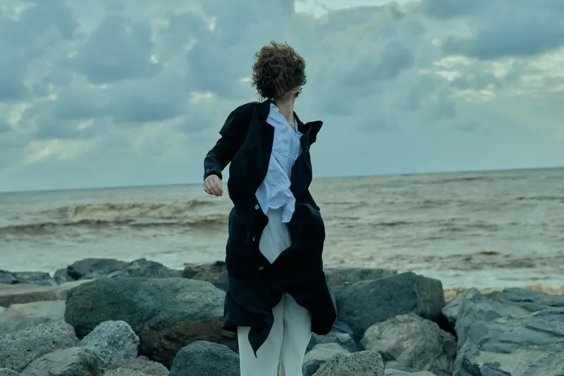 Coast scene with a coat fluttering in wind, figure on rocky shore beside a restless sea, dramatic mood and fashion inspired motion ideal for outdoorwear photography and expressive storytelling