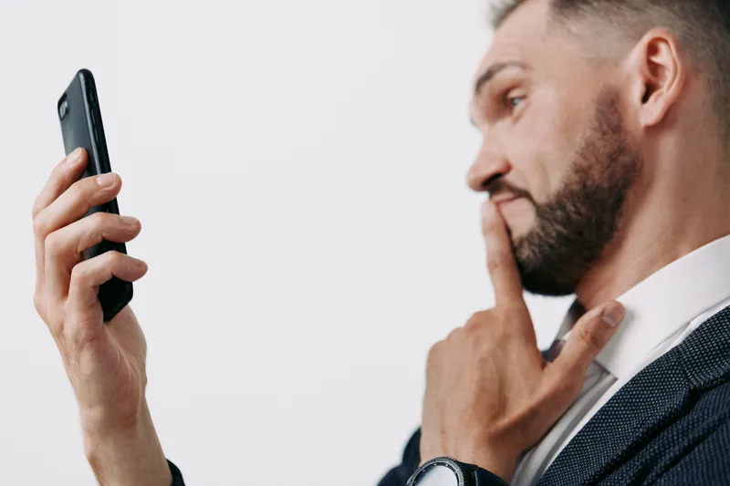 A professional man in business attire raises a thoughtful gaze toward a smartphone, standing against an isolated colorful white background, conveying focus, confidence, and ready leadership.