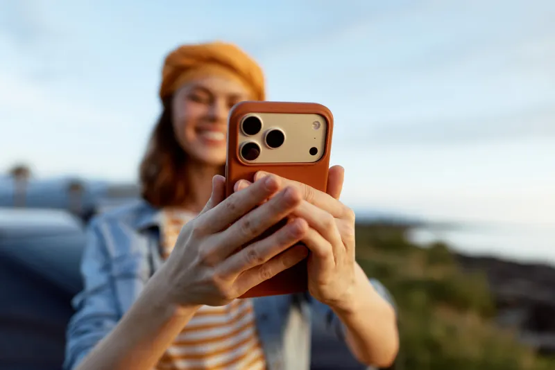 Young woman wearing a striped shirt and orange hat takes a sunny outdoor selfie, smiling into the camera while the breeze rustles her hair.