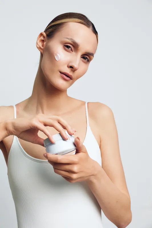 Young woman in white tank top applies face cream with fingers looking at camera isolated on gray background skin care beauty routine natural makeup healthy skin concept