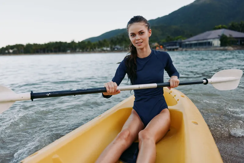 Female kayaker sits in a yellow kayak on calm water, gripping a paddle across her lap, wearing a navy swimsuit, ready for a peaceful outdoor paddle session by the shore.