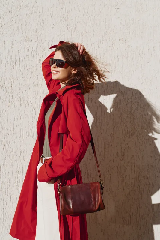 Young woman in a red coat and sunglasses stands beside a sunlit wall, adjusting her hair with a confident, stylish stride and a brown crossbody bag.