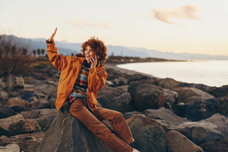 Woman lifestyle on rocky beach at sunset, wearing orange jacket and denim, curly hair blowing in sea breeze, playful pose with hand near face, coastal background with calm waves, travel and freedom