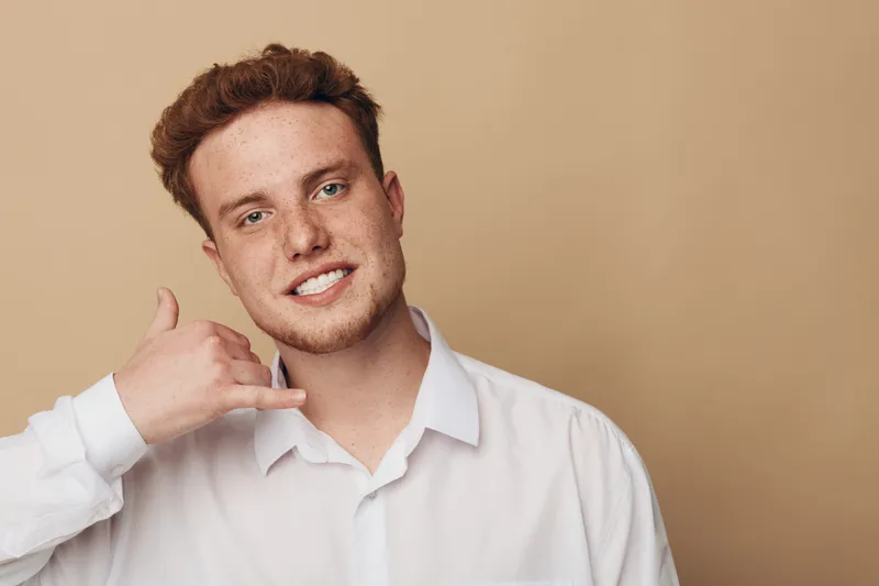 Smiling young man with red hair and freckles wearing white shirt makes call me gesture with hand near face on beige background. Concept of communication, friendliness, and casual style.