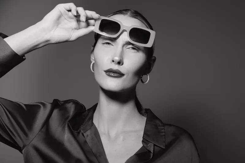 White woman with fair skin holds sunglasses near eyes, wearing dark silky blouse and hoop earrings, posing against neutral background in stylish fashion portrait.