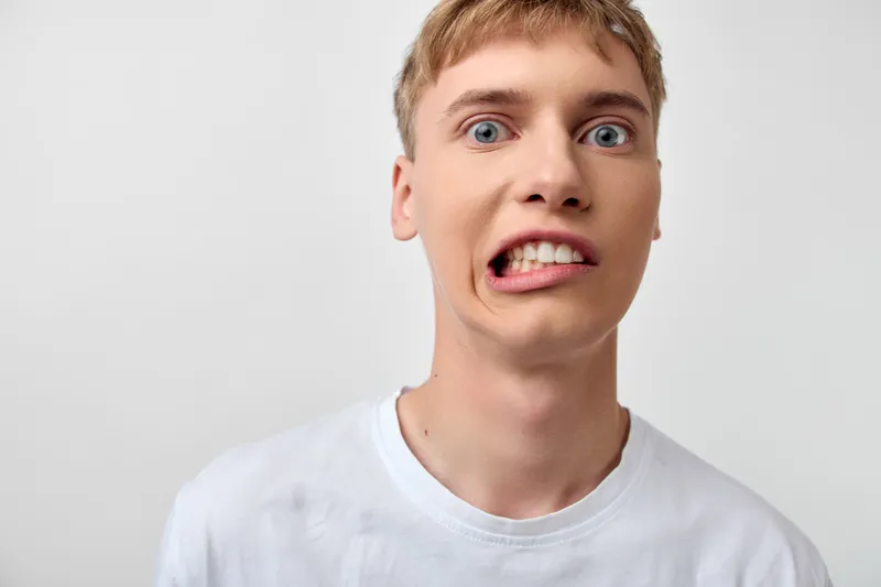 Silly face close up of a man making a goofy expression, wide eyes and stretched mouth, humorous moment perfect for comedy stock, casual white tee, plain background, playful mood with expressive