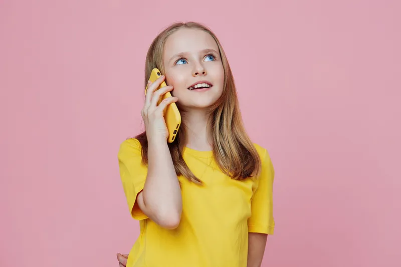 A young girl talking on the phone with a joyful expression, wearing a bright yellow shirt against a soft pink background, capturing playful childhood moments