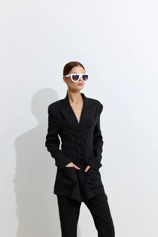 Fashion woman wearing stylish black suit and white sunglasses, posing confidently against a plain light background with hands in pockets, modern and chic look.