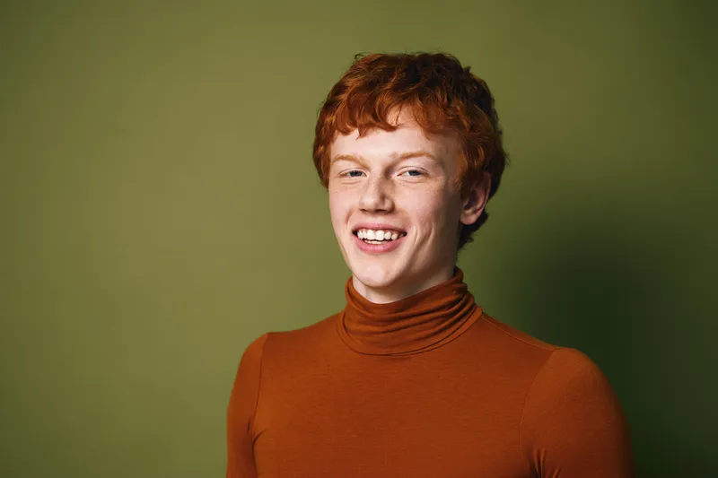 Young man with red hair wearing brown turtleneck smiling and posing against green background in studio portrait with natural light and casual style casual fashion.