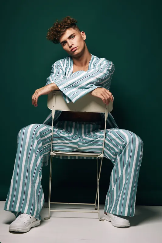 Young man in striped pajamas sitting on chair backwards with relaxed pose and curly hair against dark green background in studio.