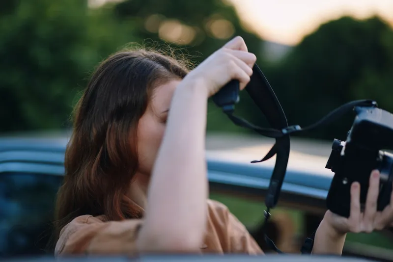 Young woman with long hair capturing memories outdoors, holding a vintage camera. Natural background. Emotions of creativity and joy.