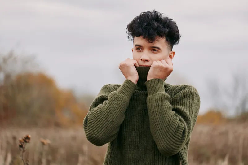 A candid outdoor portrait of a young man pulling a green knit sweater collar up around his face, capturing a genuine, realistic emotional moment with natural light and subtle detail.