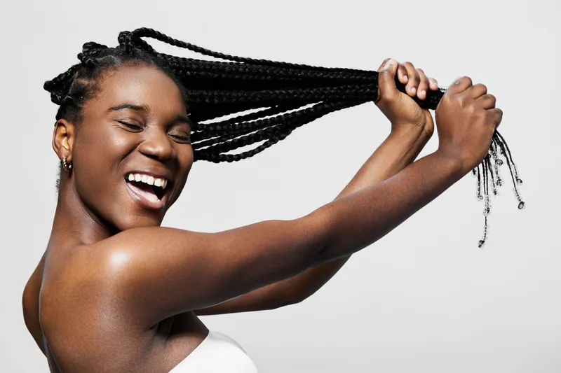 Happy young woman with braided hair smiling and playfully pulling her braids, showcasing confidence and joy in a minimalist studio background