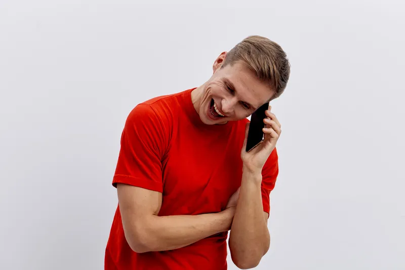 Smiling man talking on mobile phone with cheerful expression, casual red t-shirt, modern background, close-up shot, happy mood, communication concept, everyday life, leisure, lifestyle.
