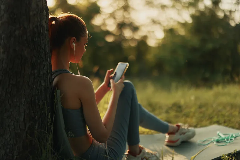 Young woman in activewear enjoying music on her phone while relaxing outdoors, showcasing a healthy lifestyle and connection with nature in a serene environment