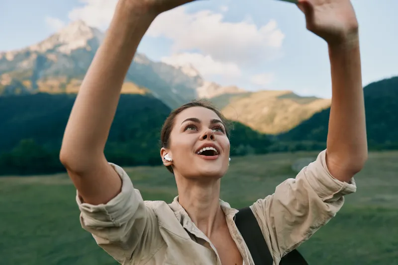 Joyful brunette woman raises her arms in a sunlit mountainside meadow, capturing a moment of outdoor adventure, freedom, and positive energy in the open air.