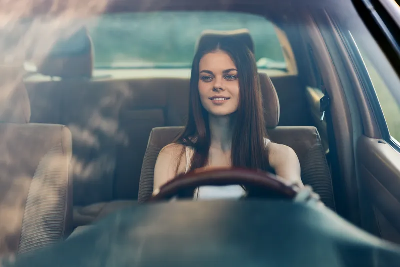 Young woman with long hair smiles while driving a car, showcasing confidence and joy during a road trip or adventure in nature.