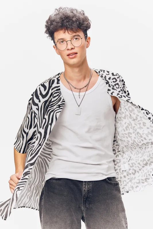 young man wearing glasses and a stylish patterned shirt posing confidently with a white tank top and casual black jeans on a white background