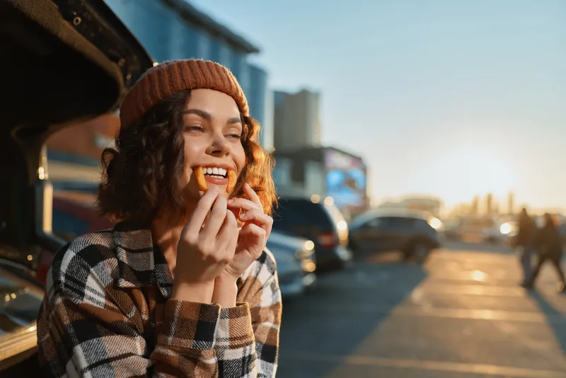 Woman smile candid lifestyle portrait in golden hour glow, authenticity and mindful living captured in an urban parking lot; beanie, curly hair and snack create emotional storytelling moment.