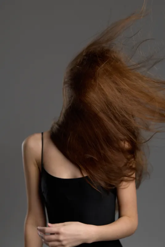 Woman with long brown hair flipping hair in motion wearing black top against gray background.