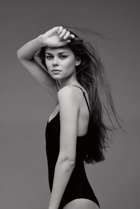 Black and white portrait of a thoughtful woman with long hair posing in a black swimsuit against a plain background.