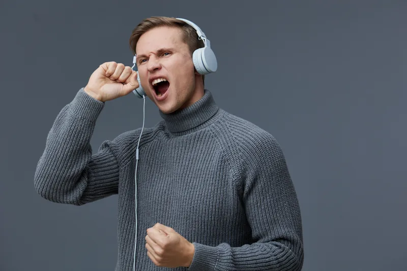 Man listening to music with headphones, expressing excitement and joy, wearing a gray sweater, against a plain gray background, studio portrait, lifestyle concept, positivity, emotional, adult male.